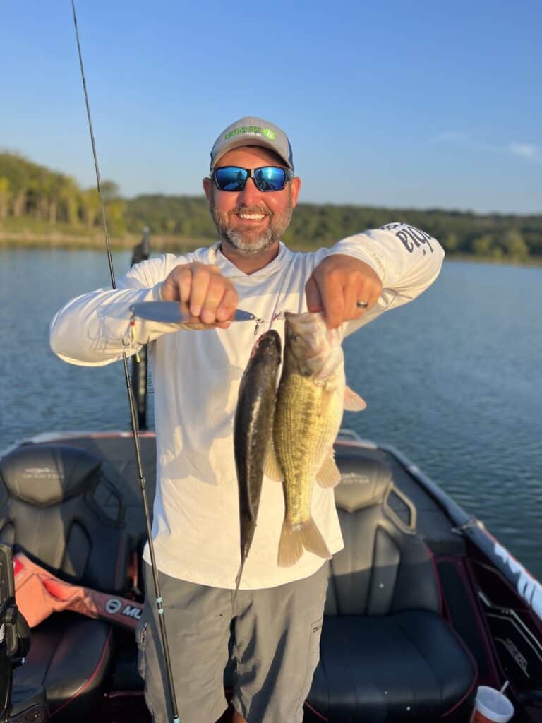 A smiling angler holds up two spotted bass he caught on the same cast while fishing at Tenkiller Lake in Oklahoma, with his boat and the lake in the background.