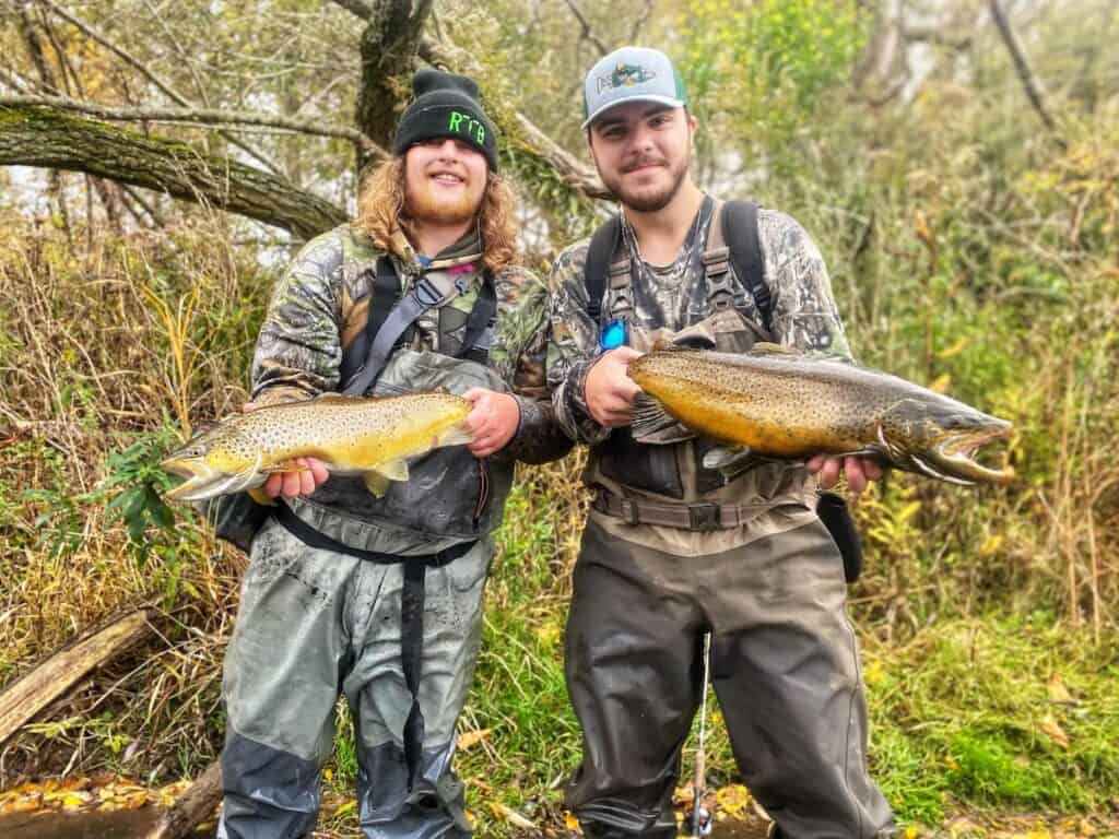 Two anglers each holding massive brown trout they caught in New York, with trees behind them.