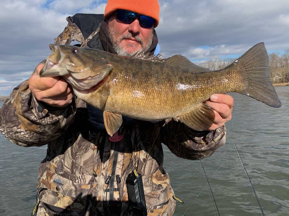 A man on a boat wearing an orange beanie and sunglasses holds up a nice smallmouth he caught fishing in the Potomac River.