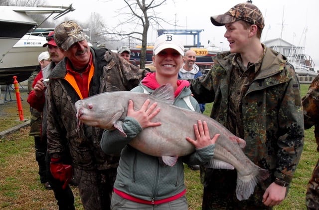 A younger angler in a white cap holds up a huge blue catfish caught in the Potomac River and two smiling men look on.