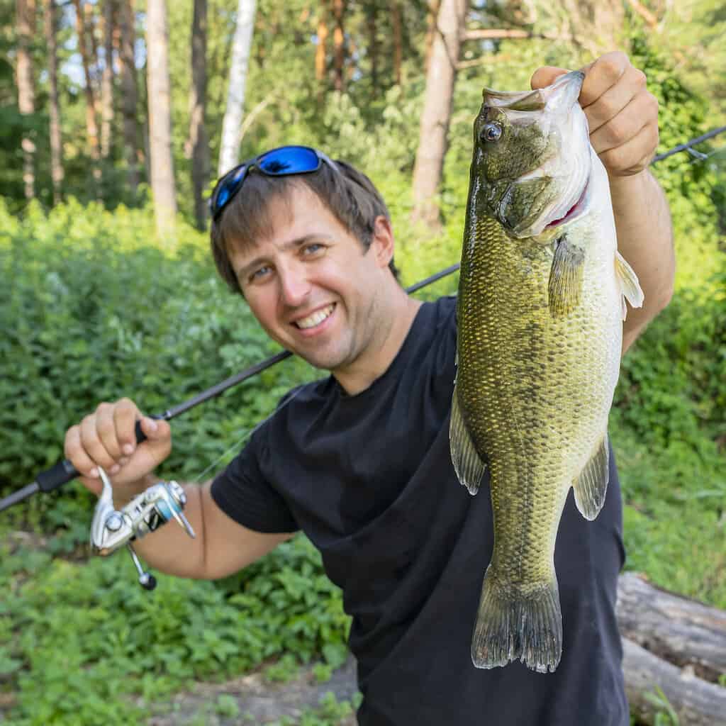 A smiling angler holds up a largemouth bass in one hand and his fishing rod in another.
